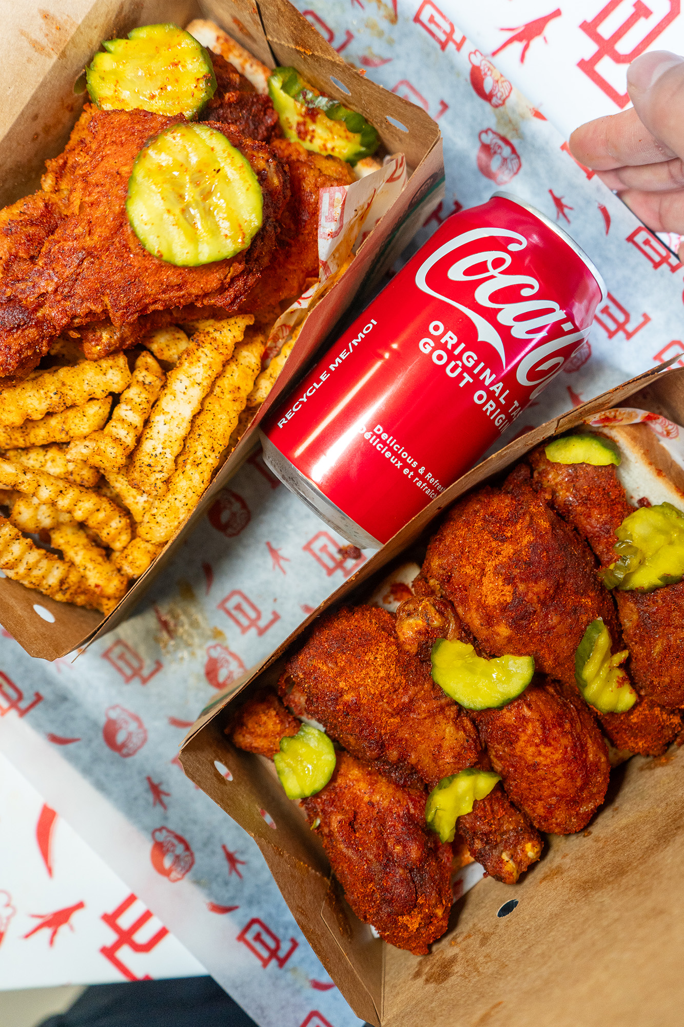 Fried drumsticks in a box, coated in a hot spice blend and topped with pickles, next to a can of coca-cola.